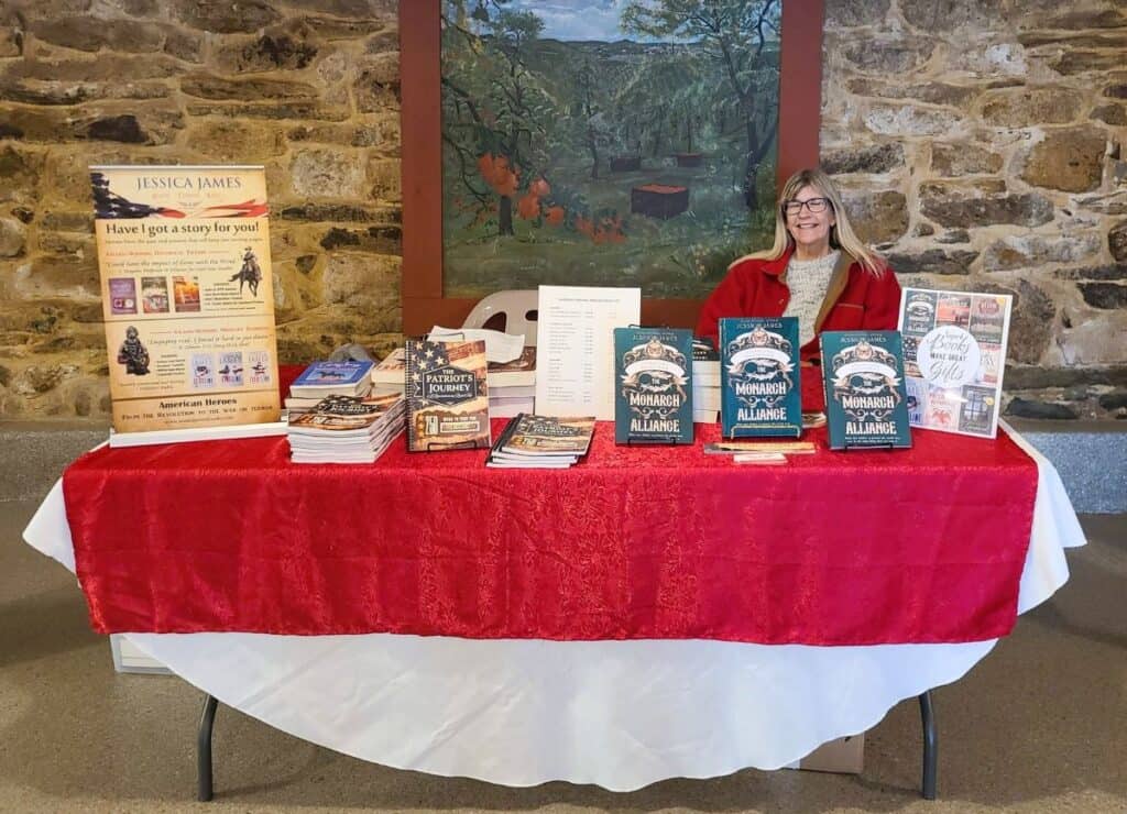 Author Jessica James wears a red coat sitting at a table with a red tablecloth with stacks of her novel The Monarch Alliance during a book signing event at the National Apple Museum.